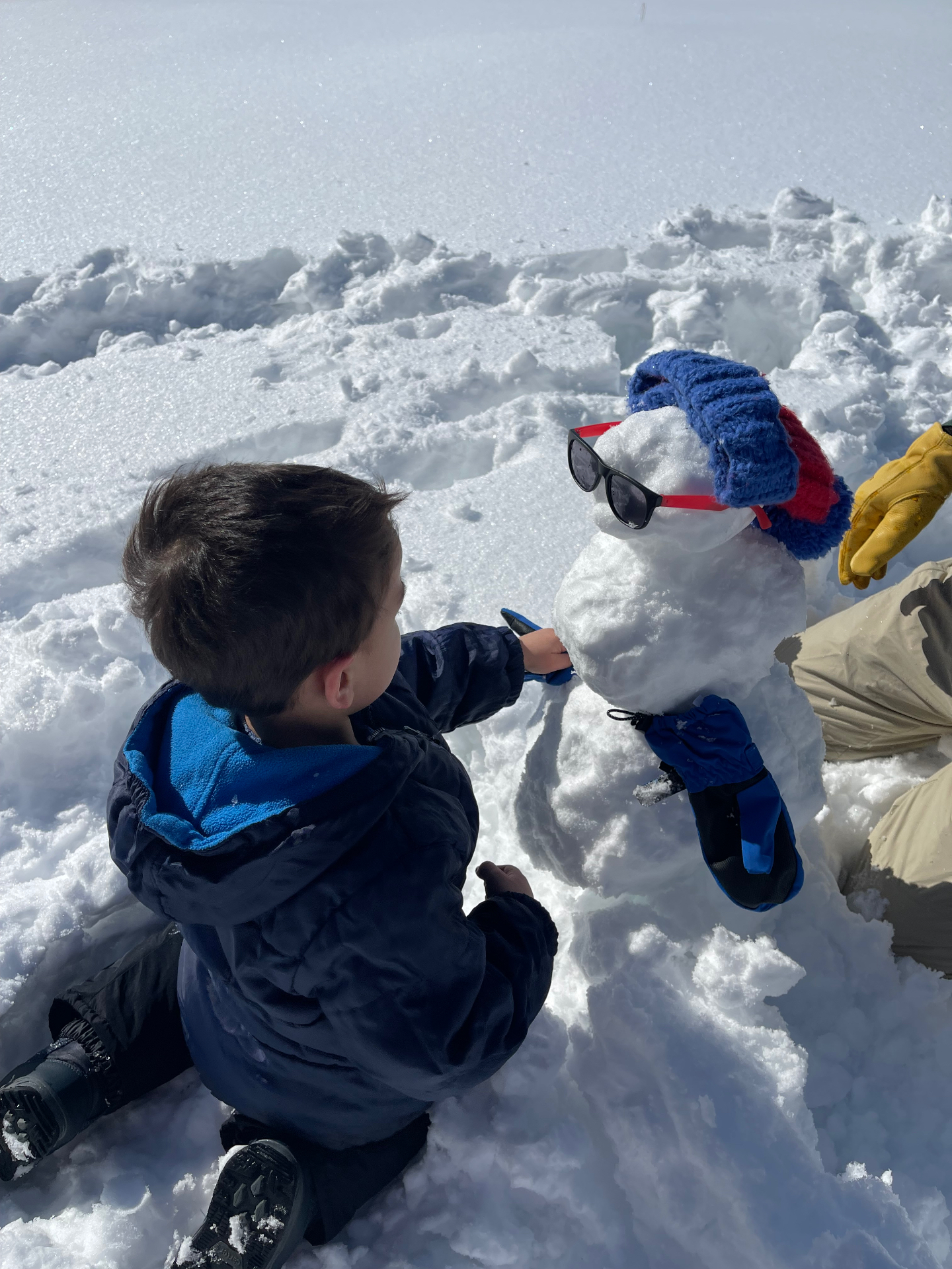 Toddler in a navy snowsuit kneeling next to a snowman wearing sunglasses and a blue and red knit hat
