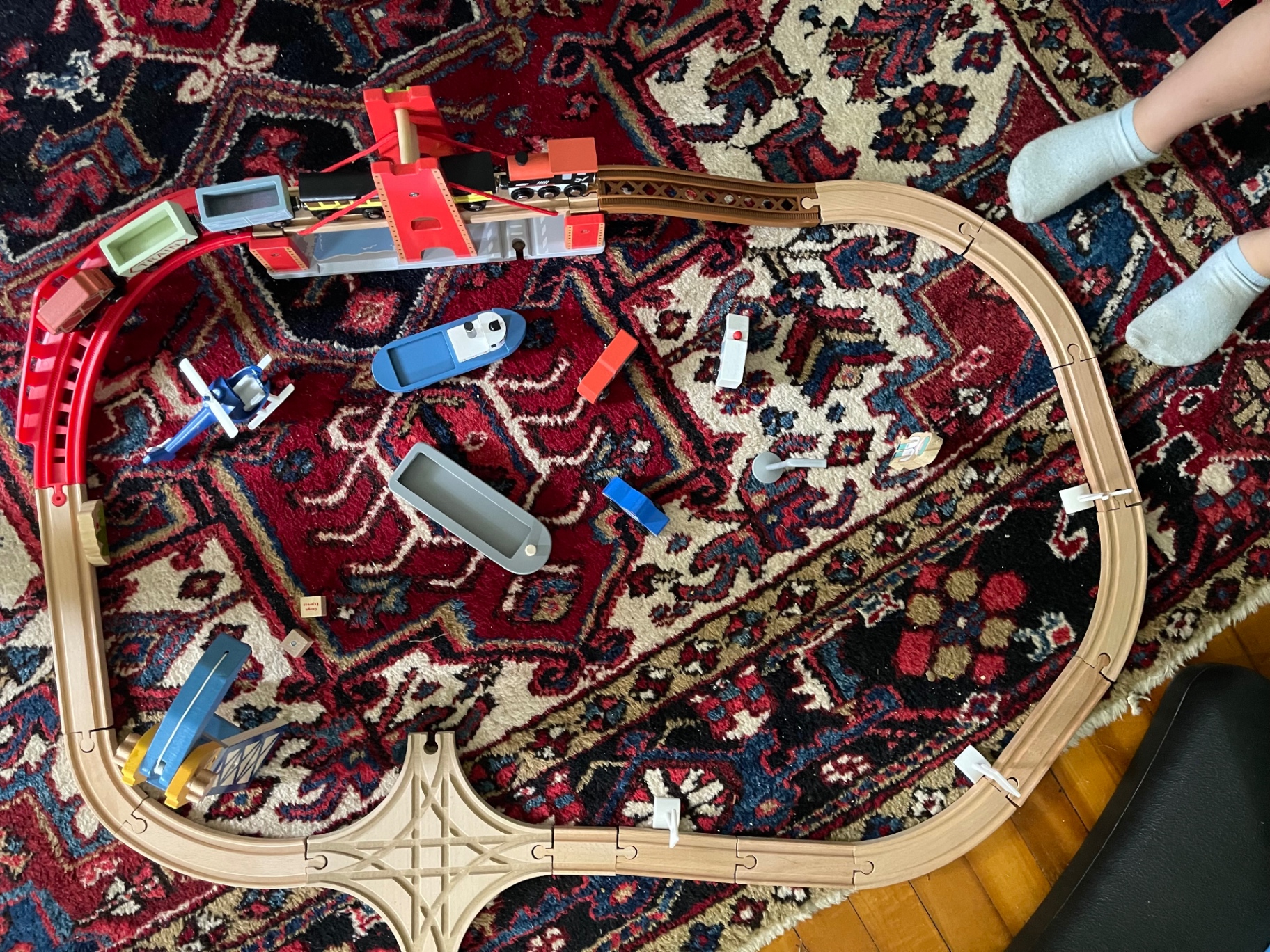 A wooden train track loop with a bridge, boats, and a helicopter laid out on a red Persian rug, small socked feet visible at the edge